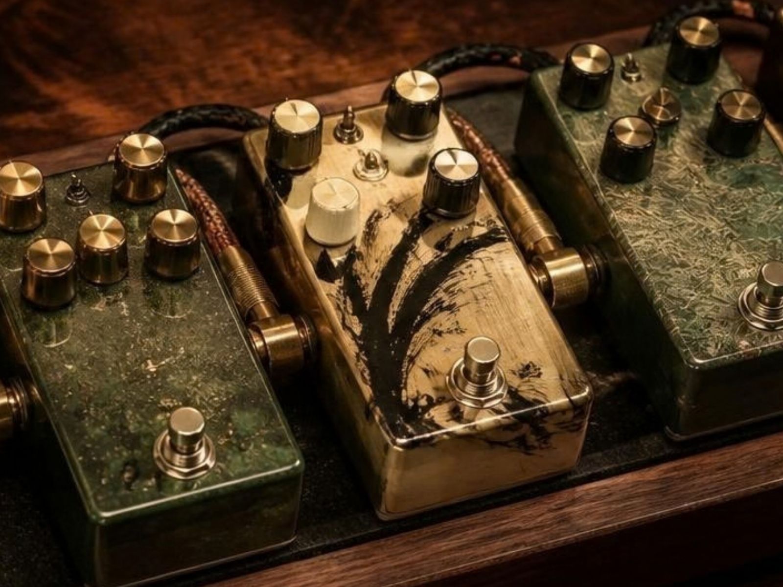 Three handmade boutique guitar pedals with brass knobs and hand-finished green and gold enclosures, linked by braided cables on a walnut bench